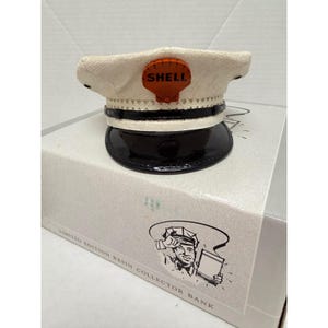 Könnte beinhalten: Eine weiße Mütze mit schwarzem Schirm und schwarzem Band, die vorne ein orangefarbenes Shell-Logo trägt. Die Mütze steht auf einer Schachtel mit der Aufschrift Limited Edition Resin Collector Bank und einer schwarz-weißen Illustration einer Person.