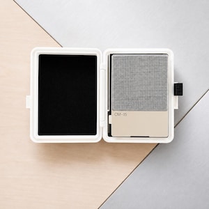 May include: A white rectangular open case with a black foam interior on the left side and a portable electronic device on the right. The device features a silver mesh speaker grille above a beige panel with the text CM-15 printed in black.