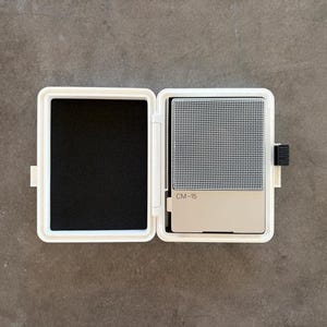 May include: A white rectangular carrying case with a hinged lid, open to reveal a black foam-lined interior on the left and a silver-toned CM-15 portable microphone device with a metallic mesh grille on the right side of the interior compartment.