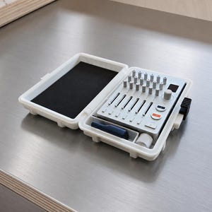 May include: A white portable hard-shell case containing a light gray audio mixing console with multiple vertical faders, rotary knobs, and a small digital display. A dark blue cylindrical tool and a small white roll are stored in the case.