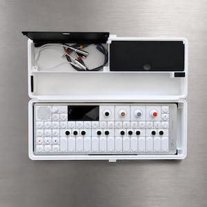 Puede incluir: Un sintetizador portátil blanco etiquetado como OP-1 con una interfaz de múltiples botones, perillas y una pantalla pequeña, alojado en una caja rectangular blanca con un compartimento de tapa negra que contiene cables de audio.