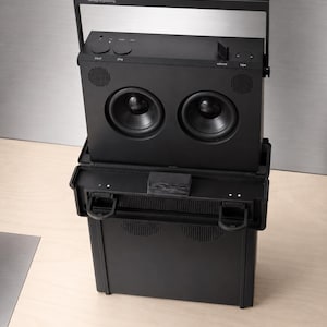 May include: A matte black portable speaker system with a rectangular frame and a top handle. The device features two prominent circular speakers, a volume dial, and control buttons labeled input, play, and tape. The text teenage engineering is printed on the top handle.