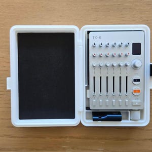 Könnte beinhalten: Weißes elektronisches Gerät mit der Aufschrift "TX-6", ausgestattet mit Knöpfen, Schiebereglern und Tasten. Es befindet sich in einem offenen weißen Gehäuse mit schwarzem Schaumstoff im Inneren. Das Gerät liegt auf einer Holzoberfläche.