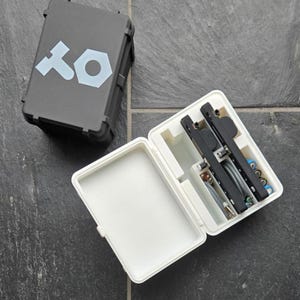 May include: A gray, rectangular case with the letters "XO" in light blue. The case is open, revealing black tools and accessories inside a white compartment. The case is on a dark gray surface.