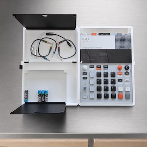 Op de afbeelding: Een witte K.O. II sampler composer met een grijs bedieningspaneel, zwarte knoppen en oranje accenten. Het apparaat staat open en toont een opbergvak met vier audiokabels en vier batterijen in de witte kunststof behuizing.