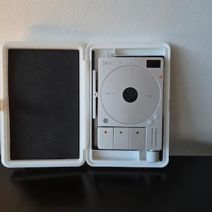 Puede incluir: Un tocadiscos portátil blanco, abierto para revelar un tocadiscos plateado con el texto "TP-2". El tocadiscos está alojado en una funda blanca con un interior negro. Se ven tres botones rojos.