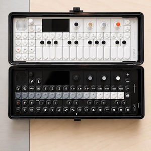 Op de afbeelding: Twee draagbare synthesizers in een zwarte draagkoffer. De bovenste unit is wit met zwarte en gekleurde knoppen, terwijl de onderste unit zwart is met grijze en zwarte toetsen. Beide apparaten hebben een toetsenbord en de tekst OP-1 erop.