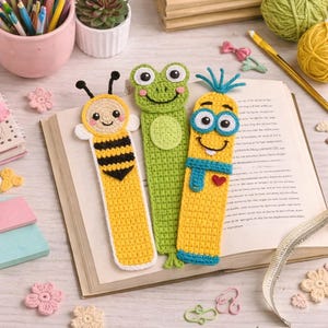May include: Three handmade crochet bookmarks: a bee, a frog, and a smiling character. The bee bookmark is yellow with black stripes, the frog is green, and the smiling character is yellow with blue accents. They are placed on an open book.