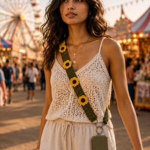 Puede incluir: Un top de ganchillo color crema y pantalones a juego se combinan con una correa verde adornada con detalles de girasoles amarillos y marrones. Una funda de teléfono está sujeta a la correa. El fondo muestra un carnaval con una noria.