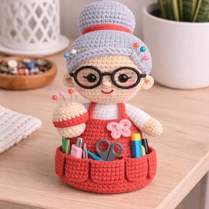 Peut inclure: Poupée crochetée représentant une femme avec des lunettes et un tablier rouge, tenant un coussin à épingles. La poupée a des cheveux gris et un visage souriant. Le tablier a des poches remplies d'outils de couture, dont des ciseaux et des stylos.