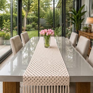 Puede incluir: Un camino de mesa rectangular de ganchillo en color crema, con un patrón de rejilla cuadrada repetitivo y acabado con largos flecos colgantes en ambos extremos. El camino está centrado sobre una mesa de comedor gris con un florero de cristal.