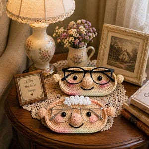 Puede incluir: Una naturaleza muerta acogedora con dos sujetagafas de ganchillo con caras sonrientes, cada uno con un par de gafas. Una foto enmarcada, una lámpara y un jarrón con flores están sobre una mesa de madera. Un letrero dice "Enjoy the little things."