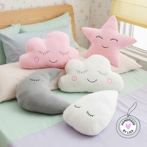 May include: A selection of plush pillows shaped like a pink star, pink cloud, white cloud, grey crescent moon, and white teardrop. Each pillow has embroidered closed eyes and a smiling mouth. The pillows are on a bed with pastel bedding.
