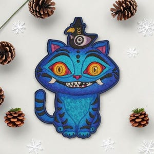 May include: A blue cat patch with a bird wearing a hat. The cat has orange eyes, sharp teeth, and a patterned body. The patch is surrounded by pine cones and snowflakes.