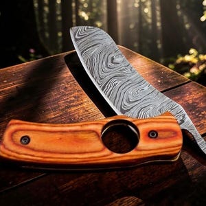 May include: A straight razor with a patterned blade rests on a wooden surface next to a wooden cigar cutter. The razor blade has a unique swirling pattern. The cigar cutter is a light brown color with a circular hole.