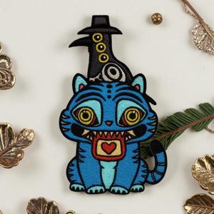 May include: Embroidered patch featuring a blue cat with yellow eyes and a red heart. A black bird-like figure with a hat sits atop the cat's head. The patch has a whimsical, cartoonish design.