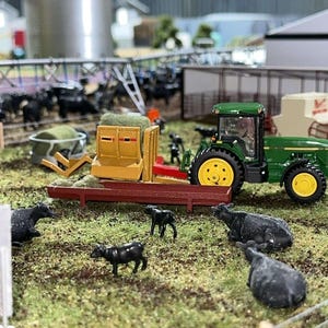 Puede incluir: Escena de granja en miniatura con un tractor verde y amarillo, un remolque de heno amarillo y varias vacas negras de juguete pastando en un campo cubierto de hierba. Un silo metálico y otras estructuras agrícolas se ven al fondo.