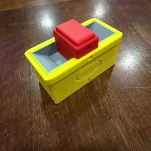 Könnte beinhalten: Eine gelbe rechteckige Box mit grauem Innenraum und einem roten rechteckigen Deckel. Die Box hat abgerundete Ecken und eine kleine, kreisförmige Vertiefung an der Seite. Der Deckel liegt auf der Box.