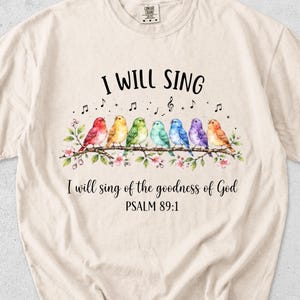 May include: An ivory short-sleeve t-shirt featuring a watercolour graphic of six colourful birds perched on a flowering branch. Above the birds is the text I WILL SING, and below is the text I will sing of the goodness of God PSALM 89:1.