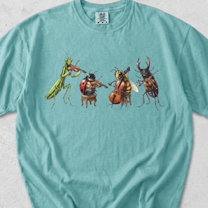 Può includere: Una t-shirt a maniche corte color verde schiuma di mare con una stampa grafica di quattro insetti che suonano strumenti musicali: una mantide religiosa con violino, una coccinella con violino, un'ape con violoncello e un cervo volante.