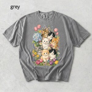 Puede incluir: Camiseta gris con una colorida ilustraci&oacute;n de gatitos entre flores. El dise&ntilde;o presenta varios gatitos con diferentes patrones de pelaje, rodeados de tulipanes rosas, hortensias azules y otras flores. La palabra "grey" est&aacute; impresa en la parte superior izquierda.
