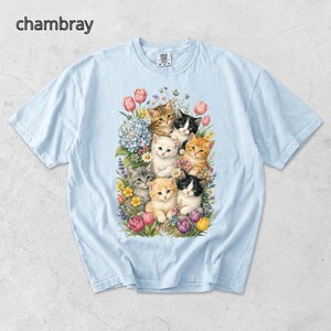 Puede incluir: Camiseta azul claro con una colorida ilustraci&oacute;n de gatitos rodeados de flores. Los gatitos tienen varios colores de pelaje, incluyendo naranja, negro y blanco. La palabra "chambray" est&aacute; impresa en la esquina superior izquierda.