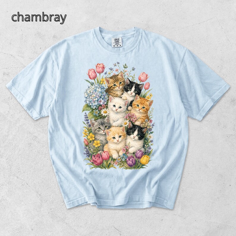Puede incluir: Camiseta azul claro con una colorida ilustraci&oacute;n de gatitos rodeados de flores. Los gatitos tienen varios colores de pelaje, incluyendo naranja, negro y blanco. La palabra "chambray" est&aacute; impresa en la esquina superior izquierda.