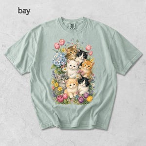 Puede incluir: Camiseta verde claro con una colorida ilustraci&oacute;n de gatitos rodeados de flores. Los gatitos tienen varios colores de pelaje, incluyendo naranja, negro, blanco y atigrado. La palabra "bay" est&aacute; impresa en la esquina superior izquierda.