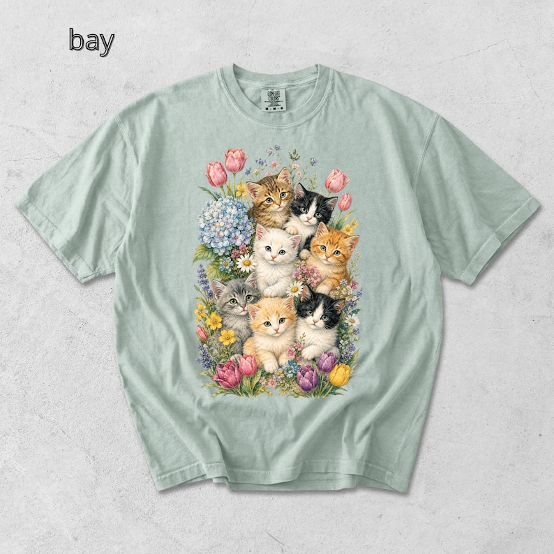 Puede incluir: Camiseta verde claro con una colorida ilustraci&oacute;n de gatitos rodeados de flores. Los gatitos tienen varios colores de pelaje, incluyendo naranja, negro, blanco y atigrado. La palabra "bay" est&aacute; impresa en la esquina superior izquierda.