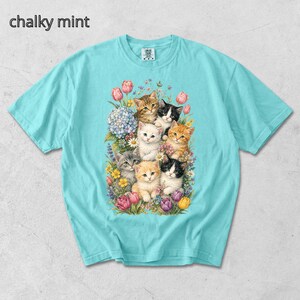 Puede incluir: Una camiseta azul claro con un gr&aacute;fico de varios gatitos rodeados de flores. El texto "chalky mint" est&aacute; en la esquina superior izquierda. Los gatitos son de varios colores, incluyendo naranja, negro y blanco.