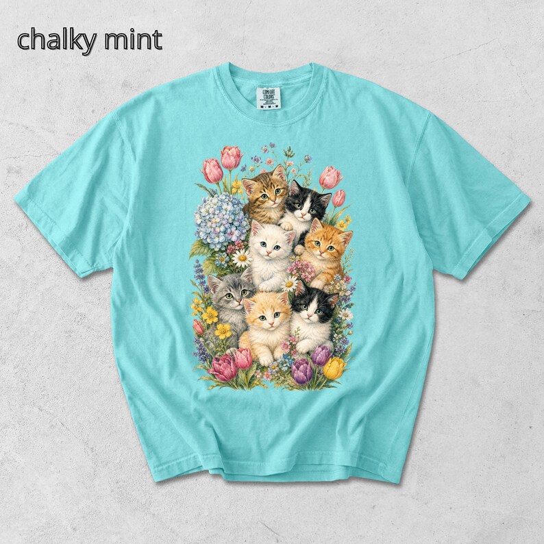 Puede incluir: Una camiseta azul claro con un gr&aacute;fico de varios gatitos rodeados de flores. El texto "chalky mint" est&aacute; en la esquina superior izquierda. Los gatitos son de varios colores, incluyendo naranja, negro y blanco.
