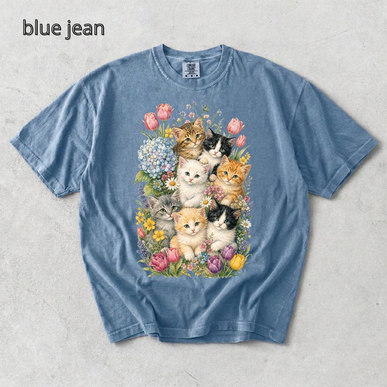 Puede incluir: Camiseta azul vaquero con una colorida ilustraci&oacute;n de gatitos rodeados de flores. Los gatitos son de varios colores, incluyendo naranja, negro y blanco. El texto "blue jean" est&aacute; en la esquina superior izquierda.