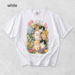 Puede incluir: Camiseta blanca con una colorida ilustraci&oacute;n de gatitos rodeados de flores. Los gatitos tienen varios colores de pelaje, incluyendo naranja, negro y gris. Las flores incluyen tulipanes, hortensias y otras flores coloridas. La palabra "white" es visible.
