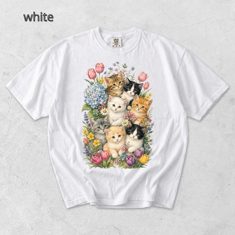 Puede incluir: Camiseta blanca con una colorida ilustraci&oacute;n de gatitos rodeados de flores. Los gatitos tienen varios colores de pelaje, incluyendo naranja, negro y gris. Las flores incluyen tulipanes, hortensias y otras flores coloridas. La palabra "white" es visible.