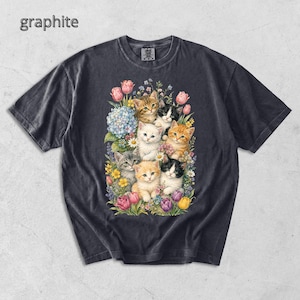 Puede incluir: Camiseta de color grafito con una colorida ilustraci&oacute;n de gatitos entre flores. El dise&ntilde;o presenta varias razas de gatos, rodeados de flores rosas, amarillas y moradas, creando una est&eacute;tica alegre y art&iacute;stica.