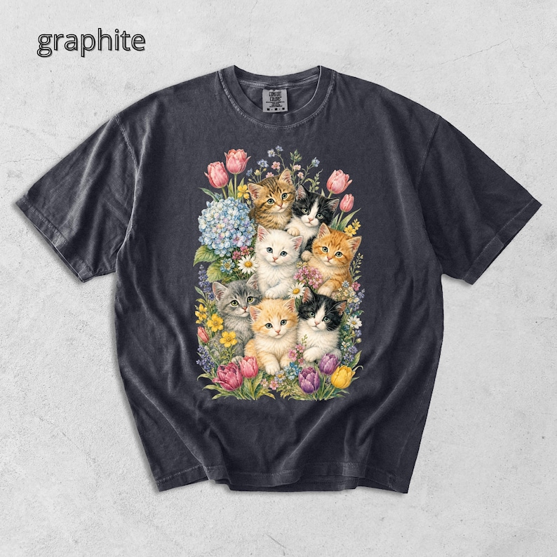 Puede incluir: Camiseta de color grafito con una colorida ilustraci&oacute;n de gatitos entre flores. El dise&ntilde;o presenta varias razas de gatos, rodeados de flores rosas, amarillas y moradas, creando una est&eacute;tica alegre y art&iacute;stica.