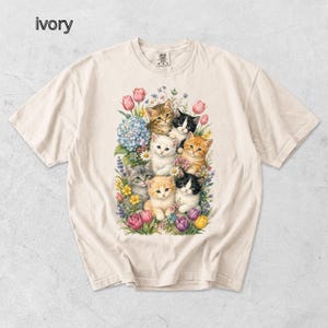 Puede incluir: Camiseta de color marfil con una ilustraci&oacute;n colorida de gatitos rodeados de flores. El dise&ntilde;o muestra un grupo de gatitos con diversos patrones de pelaje, rodeados de tulipanes rosas, hortensias azules y otras flores coloridas.