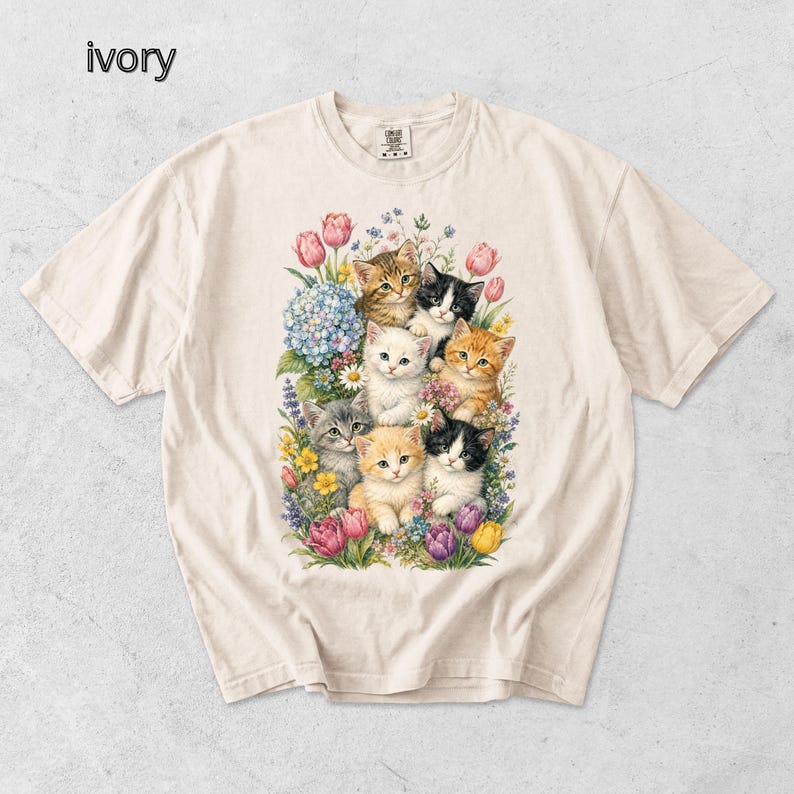 Puede incluir: Camiseta de color marfil con una ilustraci&oacute;n colorida de gatitos rodeados de flores. El dise&ntilde;o muestra un grupo de gatitos con diversos patrones de pelaje, rodeados de tulipanes rosas, hortensias azules y otras flores coloridas.