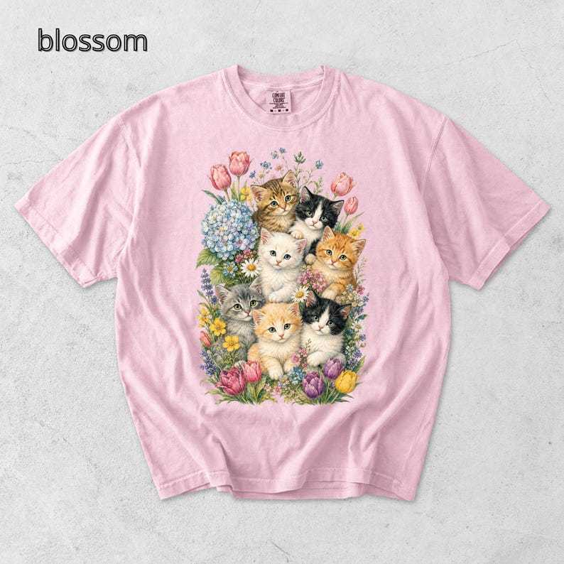 Puede incluir: Camiseta rosa claro con un estampado colorido de gatitos rodeados de flores. La palabra "blossom" est&aacute; impresa en la esquina superior izquierda. Los gatitos son de varios colores, incluyendo naranja, negro y blanco, y est&aacute;n entre tulipanes, hortensias y otras flores.