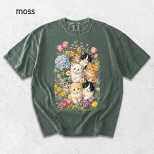 Puede incluir: Camiseta verde musgo con una colorida ilustraci&oacute;n de gatitos rodeados de flores. Los gatitos tienen varios colores de pelaje, incluyendo naranja, blanco y gris. La palabra "moss" est&aacute; impresa en la esquina superior izquierda.