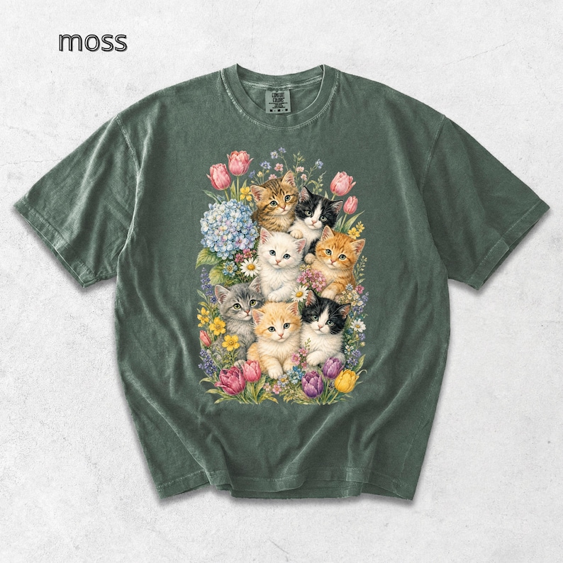 Puede incluir: Camiseta verde musgo con una colorida ilustraci&oacute;n de gatitos rodeados de flores. Los gatitos tienen varios colores de pelaje, incluyendo naranja, blanco y gris. La palabra "moss" est&aacute; impresa en la esquina superior izquierda.