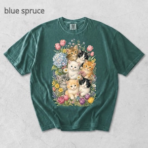 Puede incluir: Camiseta de color verde azulado con una ilustraci&oacute;n de acuarela de gatitos rodeados de flores coloridas. Los gatitos tienen varios patrones de pelaje. El texto "blue spruce" est&aacute; impreso en la parte superior izquierda.