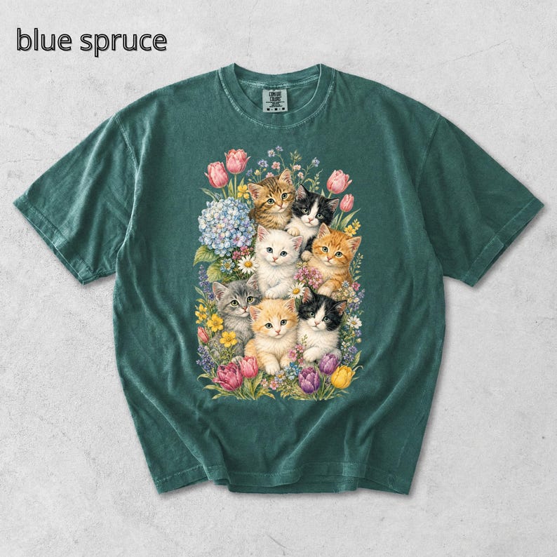 Puede incluir: Camiseta de color verde azulado con una ilustraci&oacute;n de acuarela de gatitos rodeados de flores coloridas. Los gatitos tienen varios patrones de pelaje. El texto "blue spruce" est&aacute; impreso en la parte superior izquierda.
