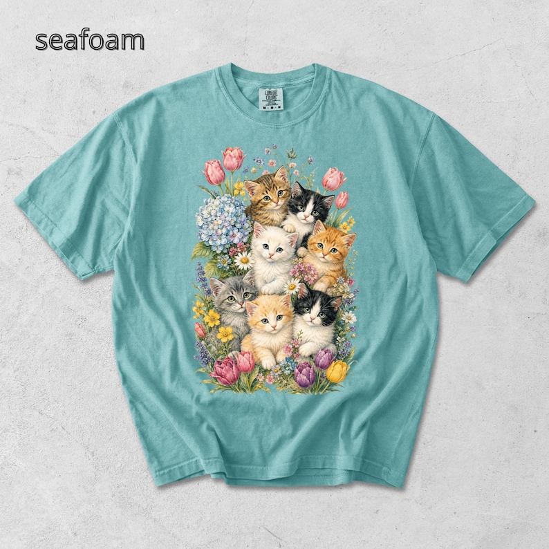 Puede incluir: Camiseta verde mar con una ilustraci&oacute;n colorida de gatitos rodeados de flores. El dise&ntilde;o muestra un grupo de gatitos con diversos patrones de pelaje, rodeados de tulipanes, hortensias y otras flores silvestres. La palabra "seafoam" est&aacute; impresa arriba a la izquierda.