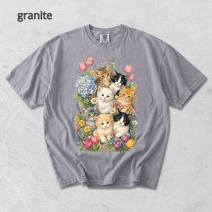 Puede incluir: Una camiseta gris con una colorida ilustraci&oacute;n de gatitos entre flores. La imagen presenta gatitos con diferentes patrones y colores de pelaje, rodeados de tulipanes rosas, hortensias azules y otras flores. La palabra "granite" est&aacute; impresa en la esquina superior izquierda.
