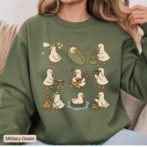 May include: Military green sweatshirt featuring a pattern of cartoon ducks engaged in various outdoor activities. The ducks are depicted with a headlamp, in a sleeping bag, using binoculars, playing guitar, and more. The text "Military Green" is at the bottom.