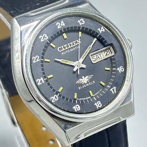 May include: A vintage Citizen automatic wristwatch with a black dial and silver-tone case. The watch features a day-date display, luminous hands and markers, and a black leather strap. The dial displays the text "CITIZEN AUTOMATIC 21 JEWELS".
