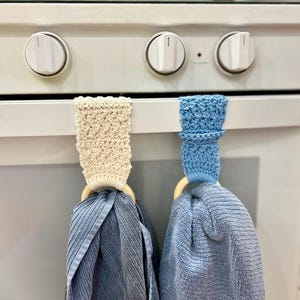 Könnte beinhalten: Zwei Küchentücher hängen an einem weißen Ofen. Eines ist hellblau, das andere dunkler blau. Jedes Tuch wird von einem Holzring und einem gehäkelten Halter gehalten, einer cremefarben und der andere hellblau.