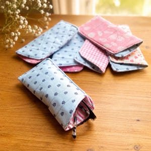 May include: A collection of fabric eyeglass cases in various patterns and colors. The cases are primarily light blue with a navy blue nautical design, and pink with floral and gingham patterns. One case holds a pair of eyeglasses.