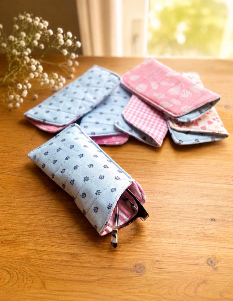 May include: A collection of fabric eyeglass cases in various patterns and colors. The cases are primarily light blue with a navy blue nautical design, and pink with floral and gingham patterns. One case holds a pair of eyeglasses.